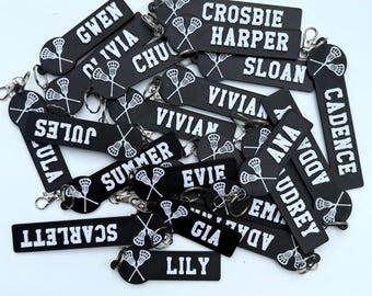 May include: A collection of black rectangular keychains featuring white text names and a white graphic of two crossed lacrosse sticks. Each keychain includes a silver metal split ring and lobster clasp attachment for securing keys.