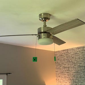 May include: A ceiling fan with silver finish and wooden blades. The fan has a light fixture and green Minecraft-themed pull chains. The fan is mounted on a white ceiling, with a brick wall in the background.