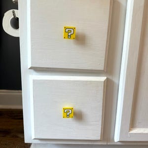 Puede incluir: Gabinete pintado de blanco con dos cajones, cada uno con un pomo amarillo y negro con un signo de interrogación. Los pomos cuadrados añaden un toque lúdico al mueble.