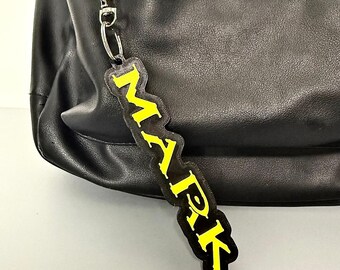 May include: A black leather bag featuring a dangling rectangular keychain tag. The tag has a black border and displays the name MARK in bold, bright yellow capital letters. The keychain is attached to the bag with a silver metal lobster clasp.