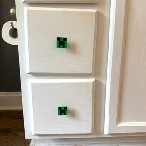 Puede incluir: Puertas de armario blancas con pomos cuadrados pixelados verdes y negros. Los pomos tienen forma de cara de Creeper de Minecraft. Las puertas del armario están pintadas de blanco con pinceladas visibles.