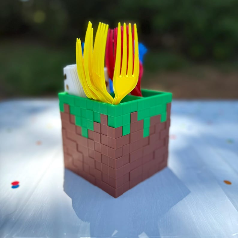 K&ouml;nnte beinhalten: Ein quadratischer, blockf&ouml;rmiger Besteckhalter mit einem braun-gr&uuml;nen Pixel-Design. Der Beh&auml;lter ist mit mehreren leuchtend gelben und roten Kunststoffgabeln gef&uuml;llt, zusammen mit einem kleinen Stapel wei&szlig;er Servietten im seitlichen Fach.
