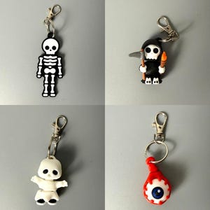 May include: Four keychains featuring spooky designs: a flat black and white skeleton, a grim reaper holding a scythe and torch, a small white ghost-like figure with black eyes, and a round red eyeball with a blue iris, all with silver clasps.