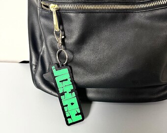 May include: A black faux leather backpack featuring a front zippered pocket with a gold-toned metal zipper. Attached to the zipper pull is a rectangular black and neon green keychain with the name JONAH printed in a bold, blocky, distressed font.