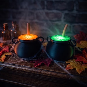 Op de afbeelding: Twee zwarte plastic ketels met handvatten, elk gevuld met een oplichtende vloeistof met bubbels. De ene ketel gloeit met een fel oranje licht, terwijl de andere een levendig groen licht uitstraalt. Uit beide steekt een lichtgekleurd rietje.
