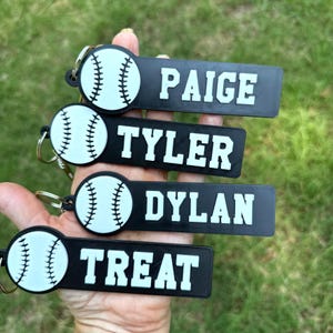 May include: Four rectangular black keychains featuring a white baseball graphic on the left side and the names PAIGE, TYLER, DYLAN, and TREAT printed in white block lettering on the right side, each attached to a silver metal split ring.