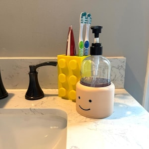 May include: A bathroom countertop scene featuring a yellow toothbrush holder, a smiling face soap dispenser, and a black faucet. The toothbrush holder is shaped like a building block. The soap dispenser has a clear bottle and a light peach base with a smiling face.