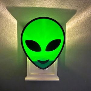 Op de afbeelding: Een groen nachtlampje in de vorm van een aliengezicht met zwarte ogen en een lachende mond. Het nachtlampje is aangesloten op een wit stopcontact en werpt een groene gloed op de muur. Het aliengezicht heeft een zwarte omtrek.