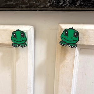 May include: Two green cartoon lizard-shaped cabinet knobs with black outlines and white eyes are attached to a white cabinet door. The knobs have a smiling mouth and a spiky head.
