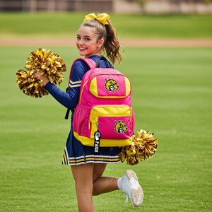 May include: A bright pink and yellow backpack featuring a mascot patch of a snarling wildcat on the front pocket and top flap. A black keychain with the name PAIGE and a pom-pom graphic hangs from the zipper, matching the gold pom-poms held.