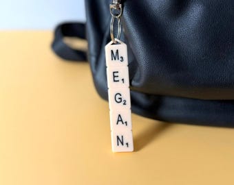 May include: A vertical keychain featuring five off-white square tiles arranged in a column, each displaying a black letter and a small numerical subscript. The tiles spell the name MEGAN and are attached to a silver-toned metal lobster clasp.