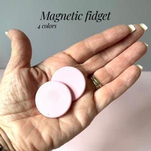 Puede incluir: Dos juguetes magnéticos fidget de color rosa claro se sostienen en una mano. Los juguetes son redondos y tienen una superficie texturizada. El texto "Magnetic fidget" y "4 colores" está en la imagen.