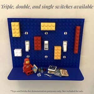 May include: A blue Lego-themed light switch plate with various colored Lego bricks and white switches. The text at the top reads "Triple, double, and single switches available." A Lego astronaut figure and miniature Lego accessories are on the shelf.