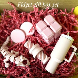 May include: A pink fidget gift box set with four colors. The box contains a variety of fidget toys, including a pink lizard, a pink twist toy, a pink tumbler, and a pink cube. The toys are nestled in red paper shreds.