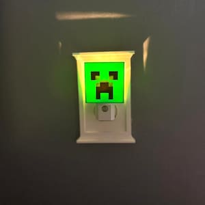 Pode incluir: Uma luz noturna branca com um design de rosto Creeper verde. O rosto Creeper é um design pixelizado com fundo verde e detalhes pretos. A luz noturna está conectada a uma tomada.