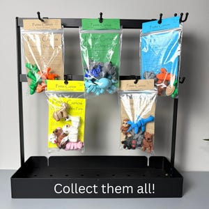 May include: A black metal display rack holding several clear plastic bags with colorful miniature figurines. The bags have labels that read "Petite Curios" and feature various themes, including dinosaurs, deep sea creatures, and farm animals. The text "Collect them all!" is at the bottom.