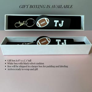May include: A black rectangular keychain featuring a brown and white football graphic and the white text TJ. The keychain includes a silver metal lobster clasp and ring, presented inside a white gift box with a black velvet cushion measuring 8 inches by 1.5 inches by 1 inch.