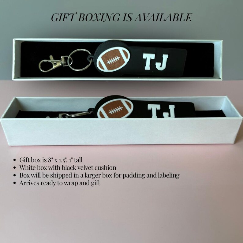May include: A black rectangular keychain featuring a brown and white football graphic and the white text TJ. The keychain includes a silver metal lobster clasp and ring, presented inside a white gift box with a black velvet cushion measuring 8 inches by 1.5 inches by 1 inch.