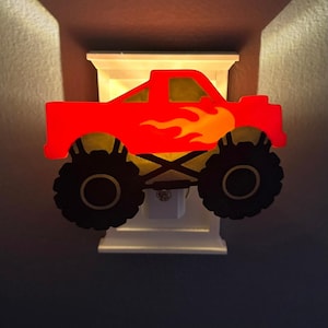 Peut inclure: Veilleuse en forme de camion monstre rouge avec des pneus noirs et un motif de flammes. La veilleuse est branchée sur une prise blanche et est montée sur une base blanche. L'intérieur du camion est jaune et éclairé.
