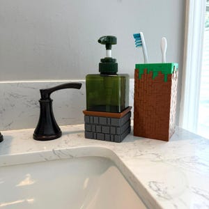 Op de afbeelding: Een groene zeepdispenser met een donkergroene pomp en een grijze en bruine blokbasis. Daarnaast een bruin-groene tandenborstelhouder in blokstijl met twee tandenborstels.