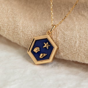 May include: Gold-toned necklace featuring a hexagonal pendant with a dark blue enamel center. The pendant is adorned with gold-colored seashell and starfish designs. The necklace is displayed on a neutral-colored fabric.