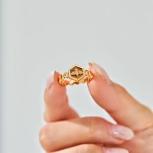 May include: A gold ring with a hexagonal bee emblem at its centre. The ring has a textured band with small, clear gemstones either side of the bee. The ring is held up against a white background.