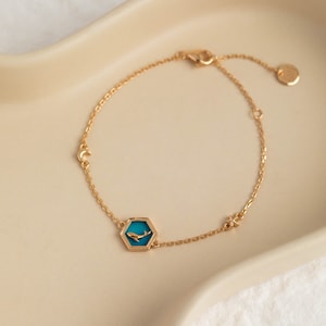 Può includere: Un braccialetto a catena dorata con un ciondolo esagonale in smalto blu che presenta una silhouette di balena. Il braccialetto ha anche un ciondolo a forma di mezzaluna e una stella.