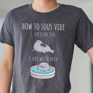 May include: Dark gray t-shirt with the text "HOW TO SOUS VIDE" and illustrations of a seal being vacuum sealed and another seal in a hot water bath. The shirt is made of a soft material.