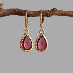 Peut inclure: Une paire de boucles d'oreilles dorées en forme de goutte d'eau, avec une bordure texturée et une pierre précieuse rouge foncé. Les boucles d'oreilles sont suspendues à une petite branche brune sur fond gris.