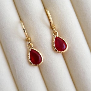 Op de afbeelding: Een paar gouden druppelvormige oorbellen met rode edelsteenaccenten. De oorbellen worden gepresenteerd in een witte fluwelen doos.