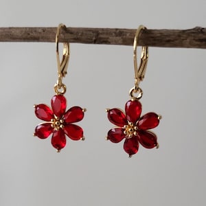 May include: A pair of gold earrings with red flower-shaped charms. Each charm has five petals and a small gold center.