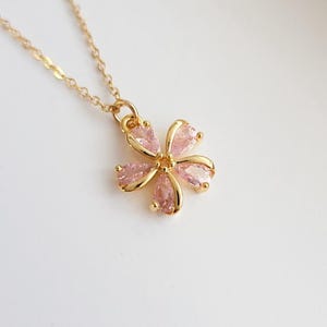 May include: A gold-tone necklace featuring a flower-shaped pendant. The pendant consists of five pear-shaped, light pink faceted stones arranged as petals, held in a gold-tone metal setting, suspended from a delicate gold-tone link chain.