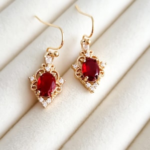 Peut inclure: Paire de boucles d'oreilles pendantes dorées avec des pierres précieuses ovales rouge foncé. Chaque boucle d'oreille est ornée de petites pierres d'accent claires et de détails en filigrane. Présentées sur une surface blanche texturée.