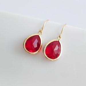 May include: A pair of drop earrings featuring faceted, pear-shaped red stones set in gold-toned metal frames. Each earring hangs from a simple gold-toned hook, showcasing a vibrant crimson hue and a polished, reflective surface on the stones.
