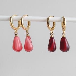 May include: Four gold hoop earrings with dangling teardrop-shaped beads. Two beads are a light pink color and two beads are a deep red color.