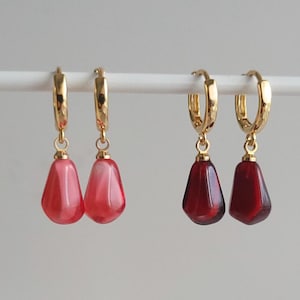 May include: Two pairs of gold hoop earrings with dangling teardrop-shaped charms. One pair has red and white swirl charms, and the other pair has solid red charms.