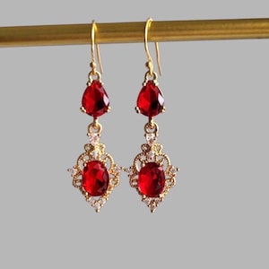 May include: A pair of gold-colored drop earrings with red teardrop and oval-shaped gemstones. The earrings are suspended from a gold-colored bar. The gemstones are surrounded by small, clear crystals.