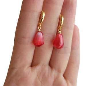 May include: A pair of gold hoop earrings with red teardrop-shaped charms. The charms have a translucent, slightly cloudy appearance.