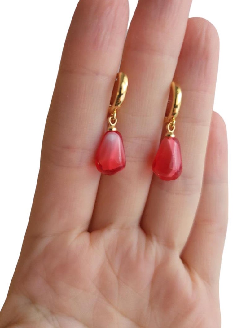 May include: A pair of gold hoop earrings with red teardrop-shaped charms. The charms have a translucent, slightly cloudy appearance.