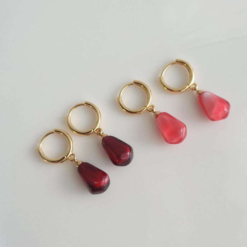 May include: Three pairs of gold hoop earrings with dangling red and pink gemstone-like drops. The earrings are on a white background.