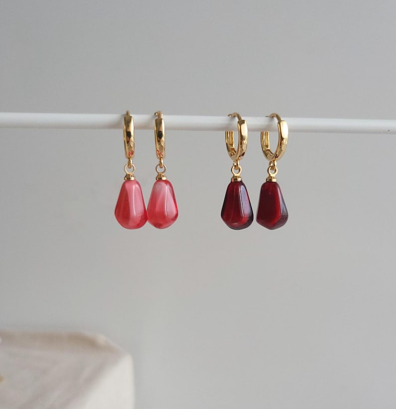 May include: Two pairs of gold hoop earrings with dangling teardrop-shaped charms. One pair has red and white swirl charms, and the other pair has solid red charms.