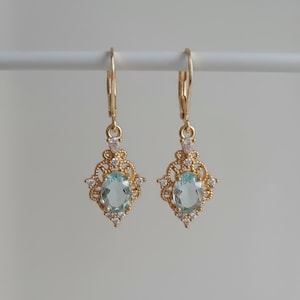 May include: A pair of gold-tone drop earrings featuring an oval-cut light blue gemstone at the center. The stone is surrounded by an intricate filigree metal frame accented with small clear stones and suspended from a lever-back closure.