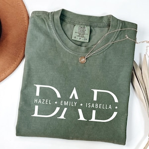 May include: An olive green t-shirt with the word "DAD" in large white letters. Below the word "DAD" are the names "HAZEL", "EMILY", and "ISABELLA". A gold necklace with a pendant rests on the shirt. A brown hat is in the background.
