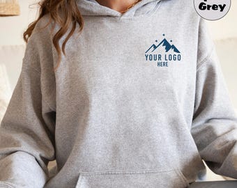 Puede incluir: Una sudadera con capucha gris con un bolsillo y un pequeño gráfico de cordillera con el texto "YOUR LOGO HERE" en la parte delantera.
