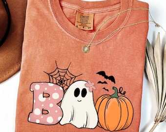 Puede incluir: Camiseta naranja Comfort Colors con un diseño de Halloween. El diseño incluye la palabra "BOO", un fantasma, una telaraña, murciélagos y una calabaza. Un collar dorado está sobre la camiseta.