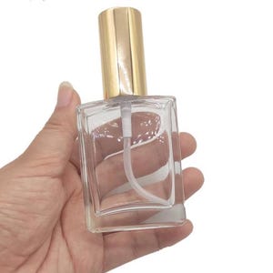 May include: A clear, rectangular glass perfume bottle with a gold-colored cap and a white spray nozzle. The bottle is held in a hand, showcasing its shape and design. The bottle is empty.