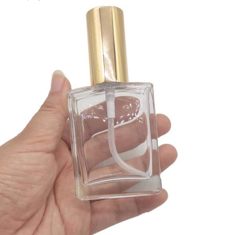 May include: A clear, rectangular glass perfume bottle with a gold-colored cap and a white spray nozzle. The bottle is held in a hand, showcasing its shape and design. The bottle is empty.