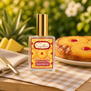 Puede incluir: Un frasco de perfume transparente con tapa dorada y una etiqueta con rodajas de piña y el texto "Sunny Side Up". El frasco está junto a un pastel de piña invertido y un cuchillo sobre un mantel a cuadros.