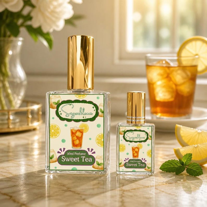 May include: Two glass perfume bottles with gold caps, labeled "Sugarbloom Sweet Tea." The labels feature a sweet tea illustration, lemons, and peaches. A glass of iced tea with a lemon slice and mint leaves are in the background.