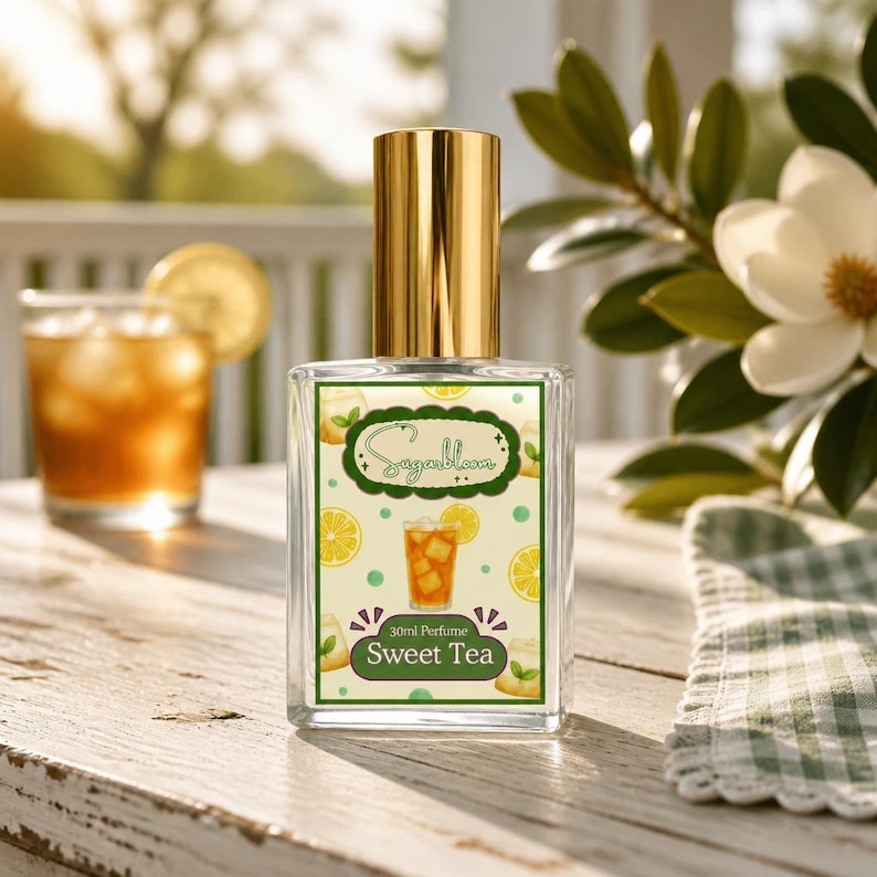 May include: A clear glass perfume bottle with a gold cap, labeled "Sugarbloom 30ml Perfume Sweet Tea." The label features a graphic of iced tea with lemon slices and lemon illustrations. The bottle sits on a wooden table with a glass of iced tea in the background.